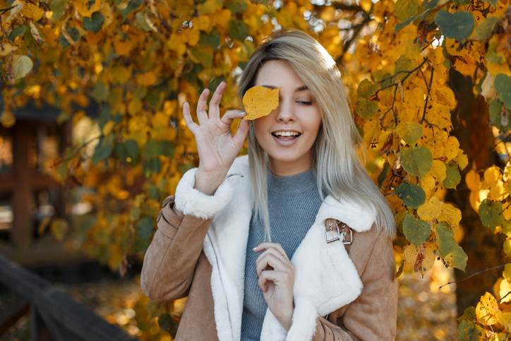 Pretty young happy girl model with blue eyes in a fashion jacket with fur covered her face with a yellow autumn leaf in the gold fall park . Beautiful smiling woman.