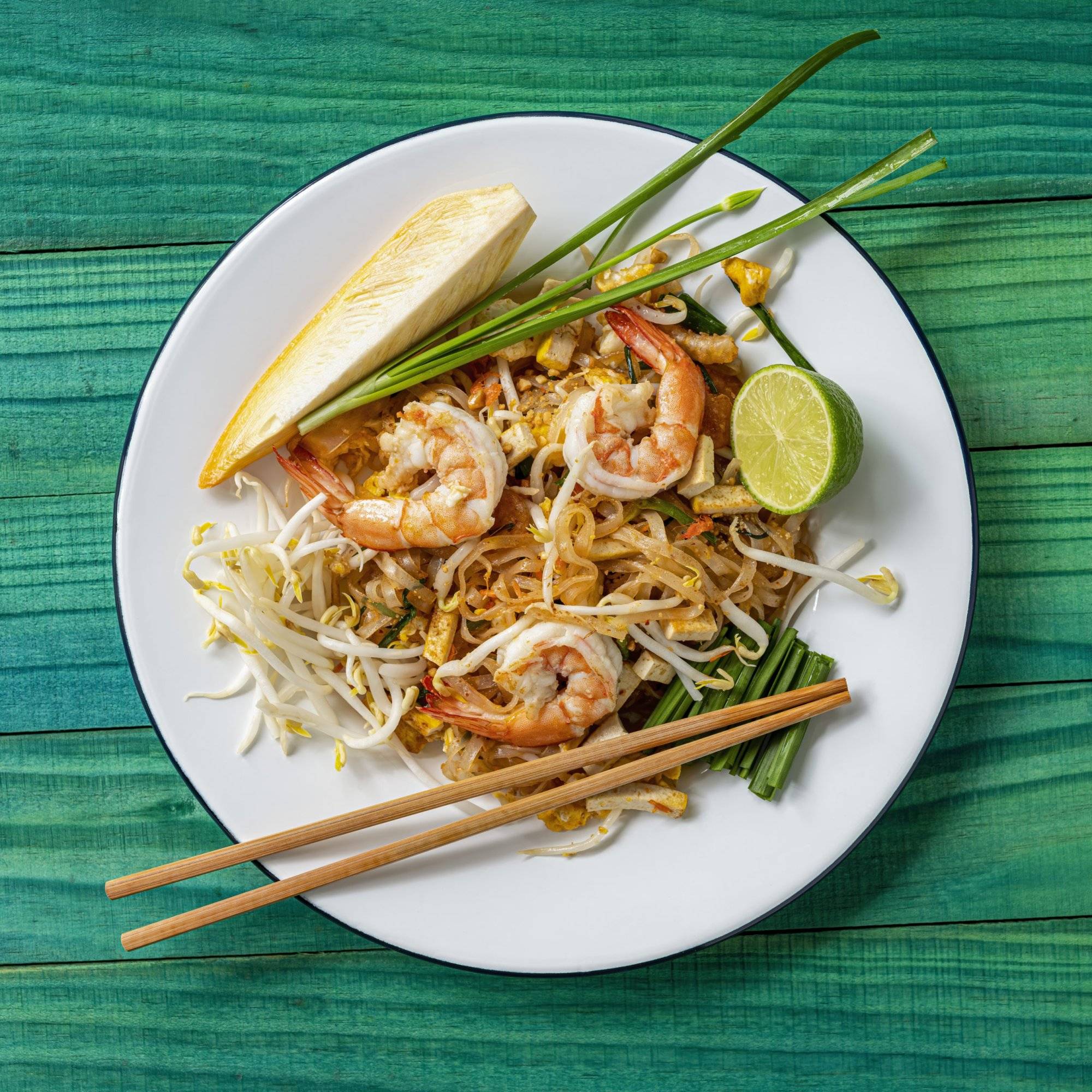 World famous freshly stir-fried cooked, Thai recipe of Prawn Pad Thai noodles on a round traditional enameled metal dish with chopsticks laid on the side of the dish, set on an abstract weathered turquoise colored wood panel table background.